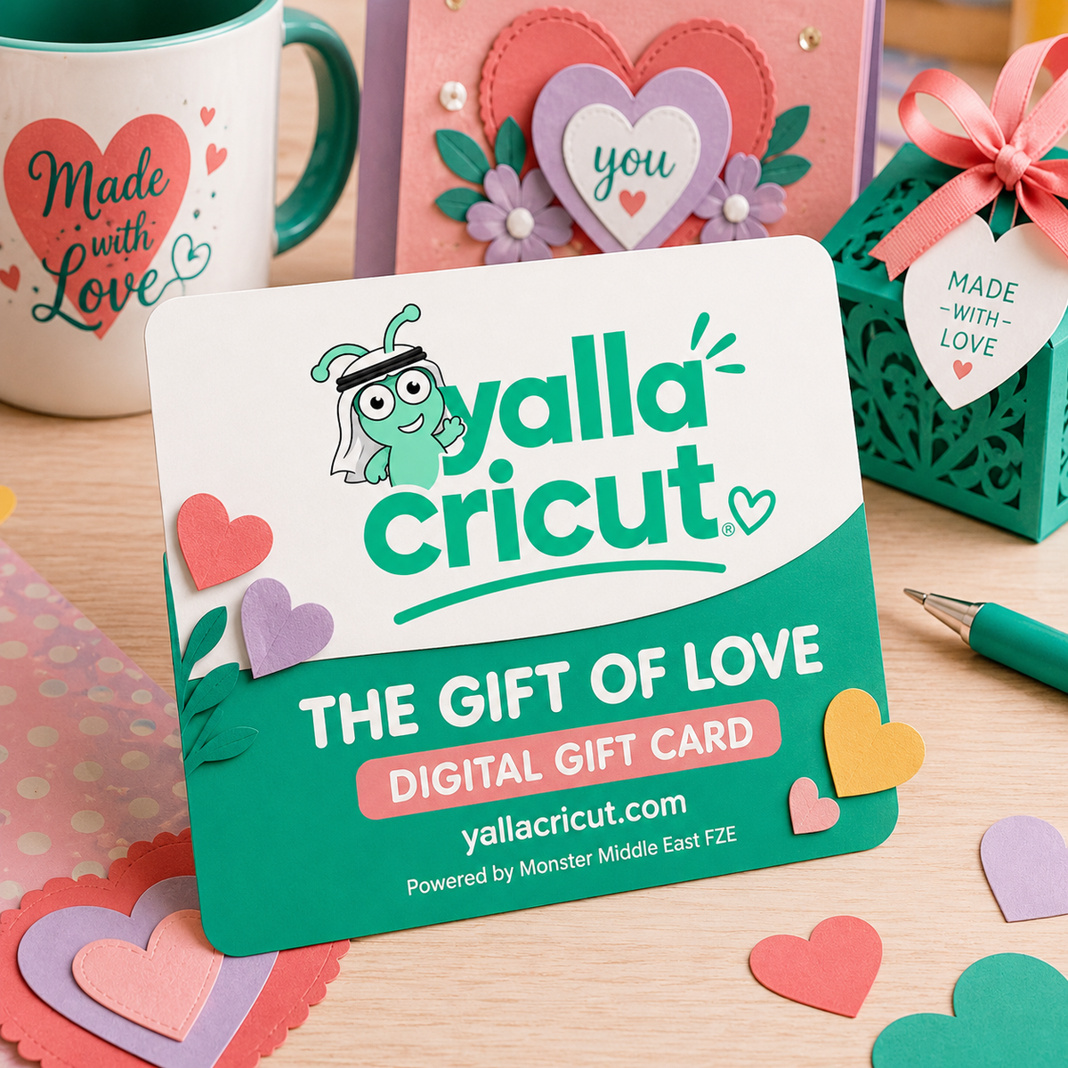 A Yalla Cricut digital gift card standing on a wooden table, surrounded by heart-shaped paper cutouts and other gift items in the background. The card is white and teal with the Yalla Cricut logo and text reading 'The Gift of Love Digital Gift Card'.